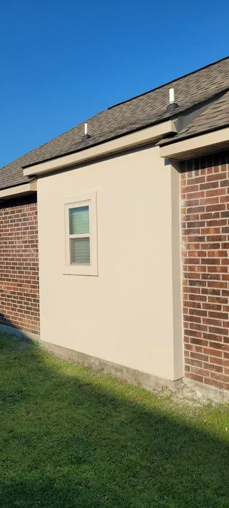 A tan stucco wall with a small window between brick walls, with a roof and green grass in the foreground.