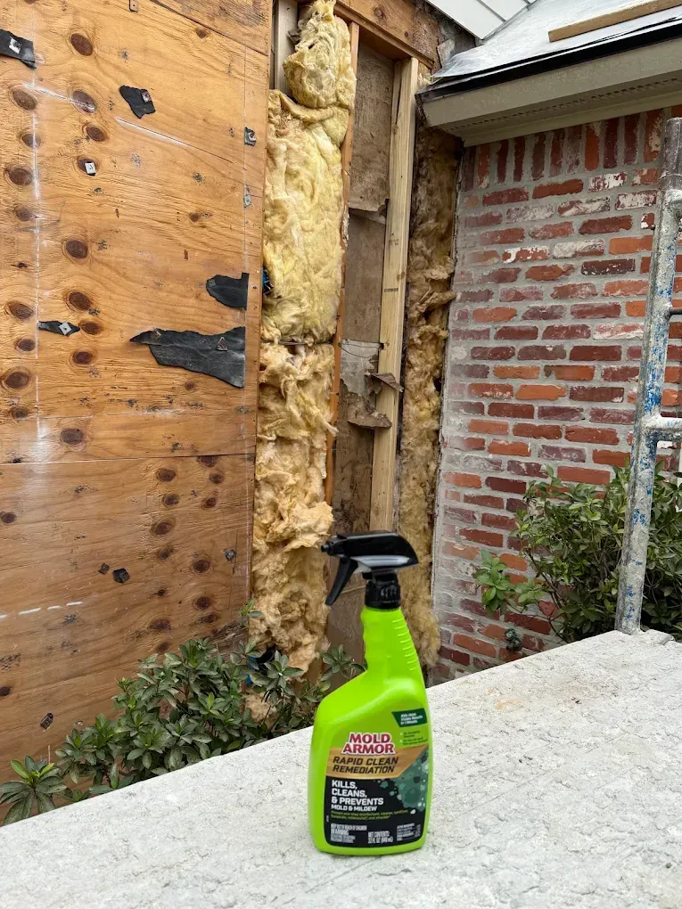 Green spray bottle in front of exposed wall insulation, brick wall, and plywood.