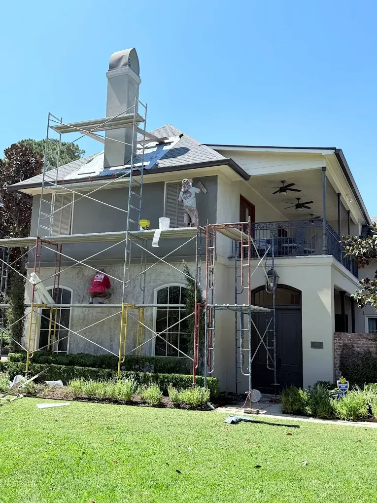 House exterior being renovated; scaffolding, workers, gray stucco, and blue sky.
