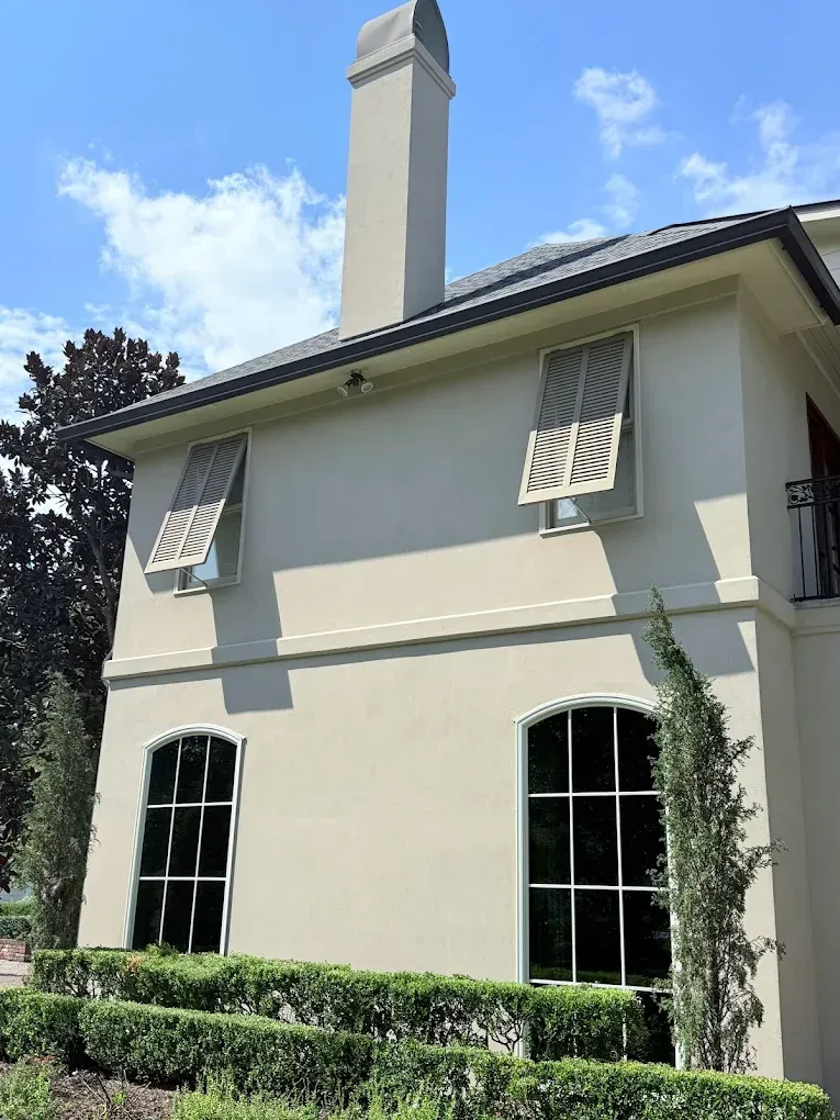 Beige stucco building with shutters, chimney, and large windows against a blue sky.