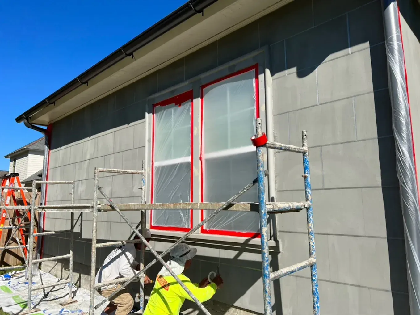 Exterior house renovation: worker on scaffolding applies coating to siding, window covered.