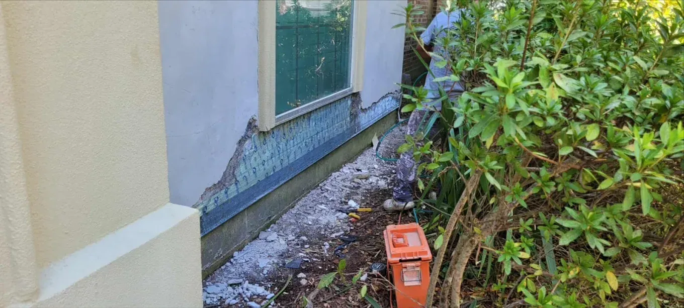 Damage to the siding and foundation of a building is visible. A person is near overgrown bushes.
