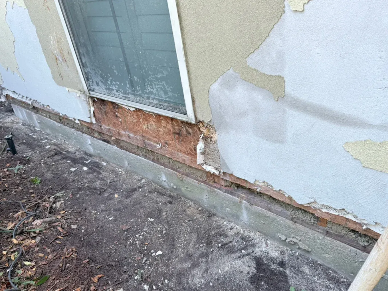 Exterior wall with peeling stucco, exposed wood near window, and concrete foundation.