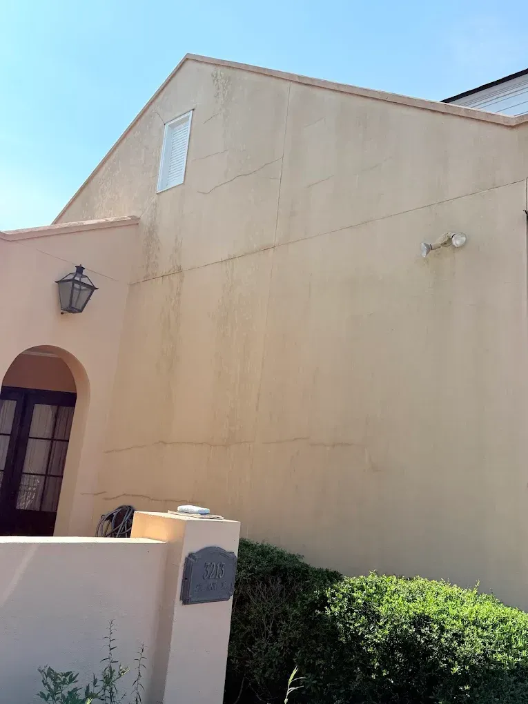 Beige stucco building with arched entrance, small window, security camera, and lush greenery.