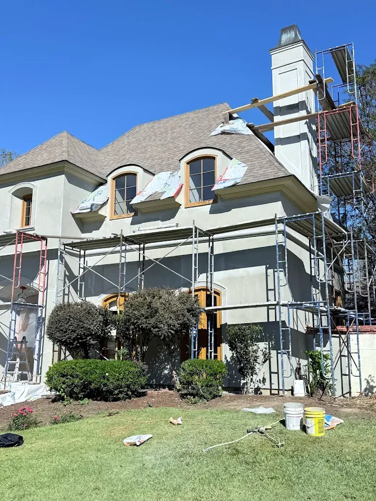 House exterior under renovation with scaffolding; beige stucco walls, brown roof, and blue sky.