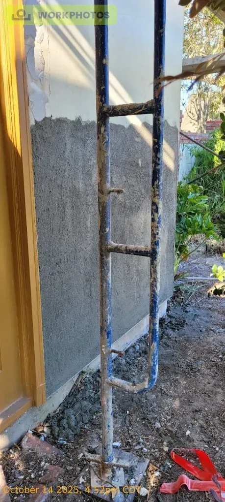 A blue metal scaffolding next to a wall with gray stucco. Ground is dirt and debris.