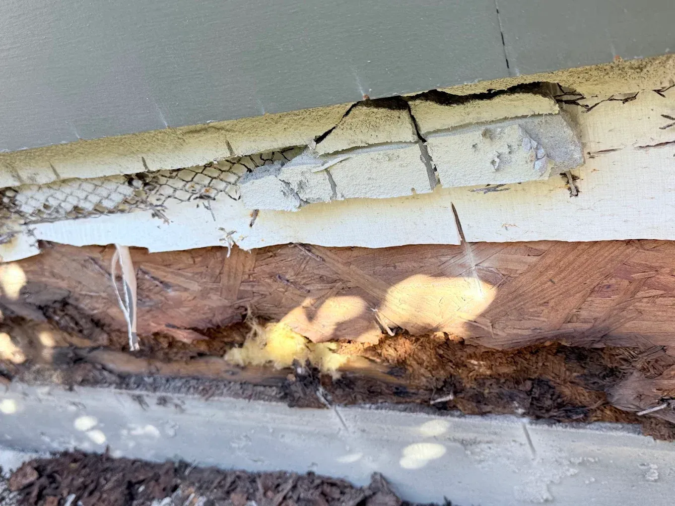 Damaged exterior wall showing crumbling stucco, exposed wood, and metal mesh.
