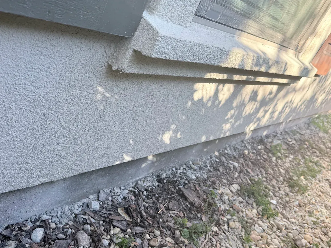 Exterior house wall with textured light grey stucco, below a window and cement foundation.