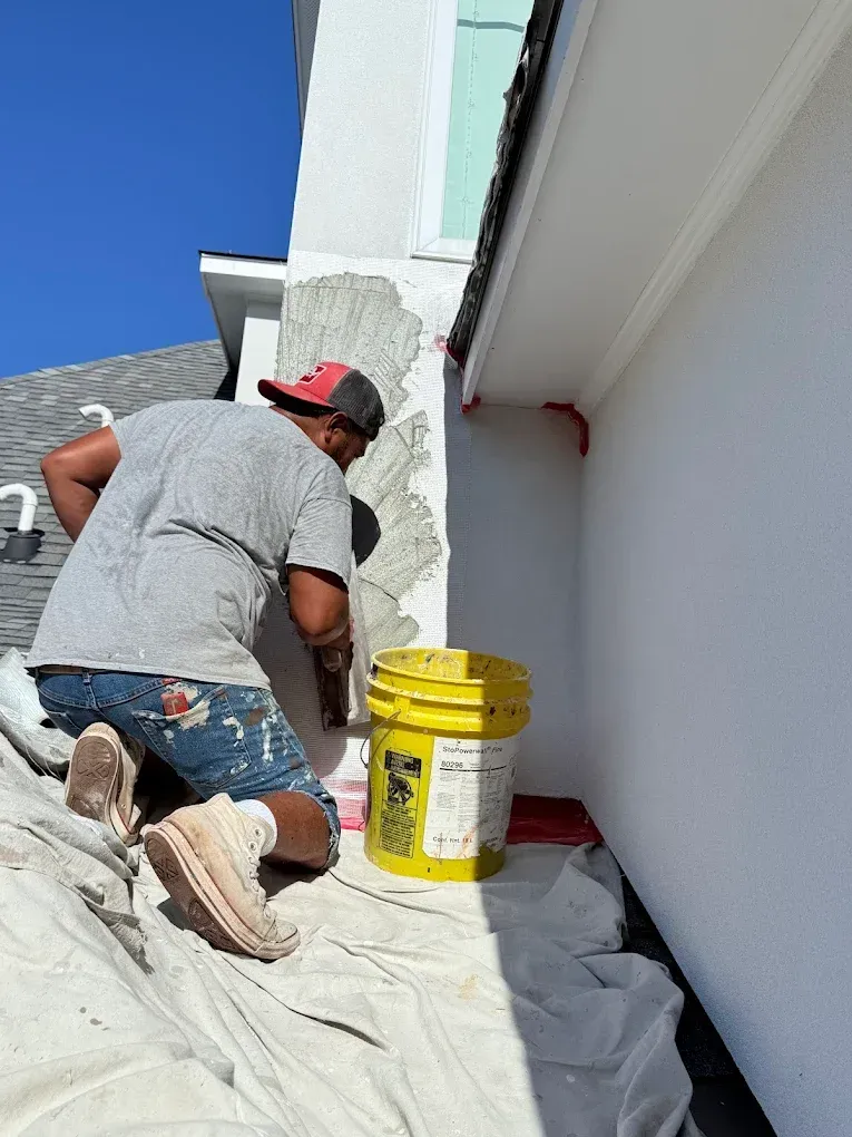 Person applying stucco to exterior wall corner, with bucket, tarp, and blue sky.
