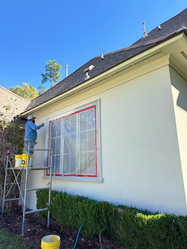 A person painting the exterior wall of a house, standing on a ladder. The window is covered with plastic.