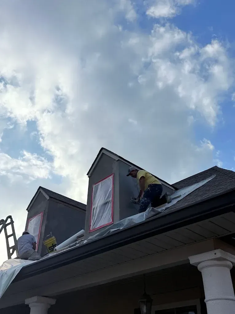 Workers on a rooftop, repairing a gray dormer under a cloudy sky.