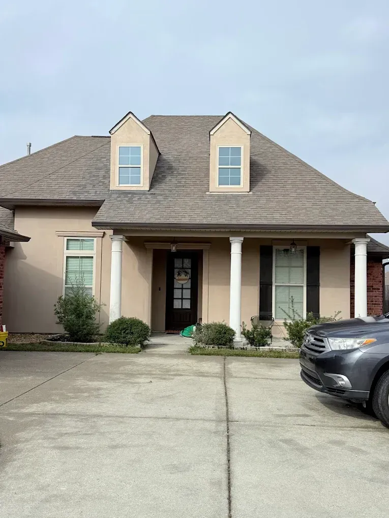 Beige stucco house with brown roof and two dormers. White columns support a covered porch with a dark front door.