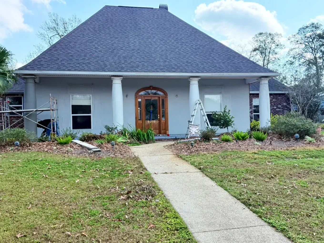 Gray house with columns and brown front door, surrounded by lawn and garden. Construction equipment visible.