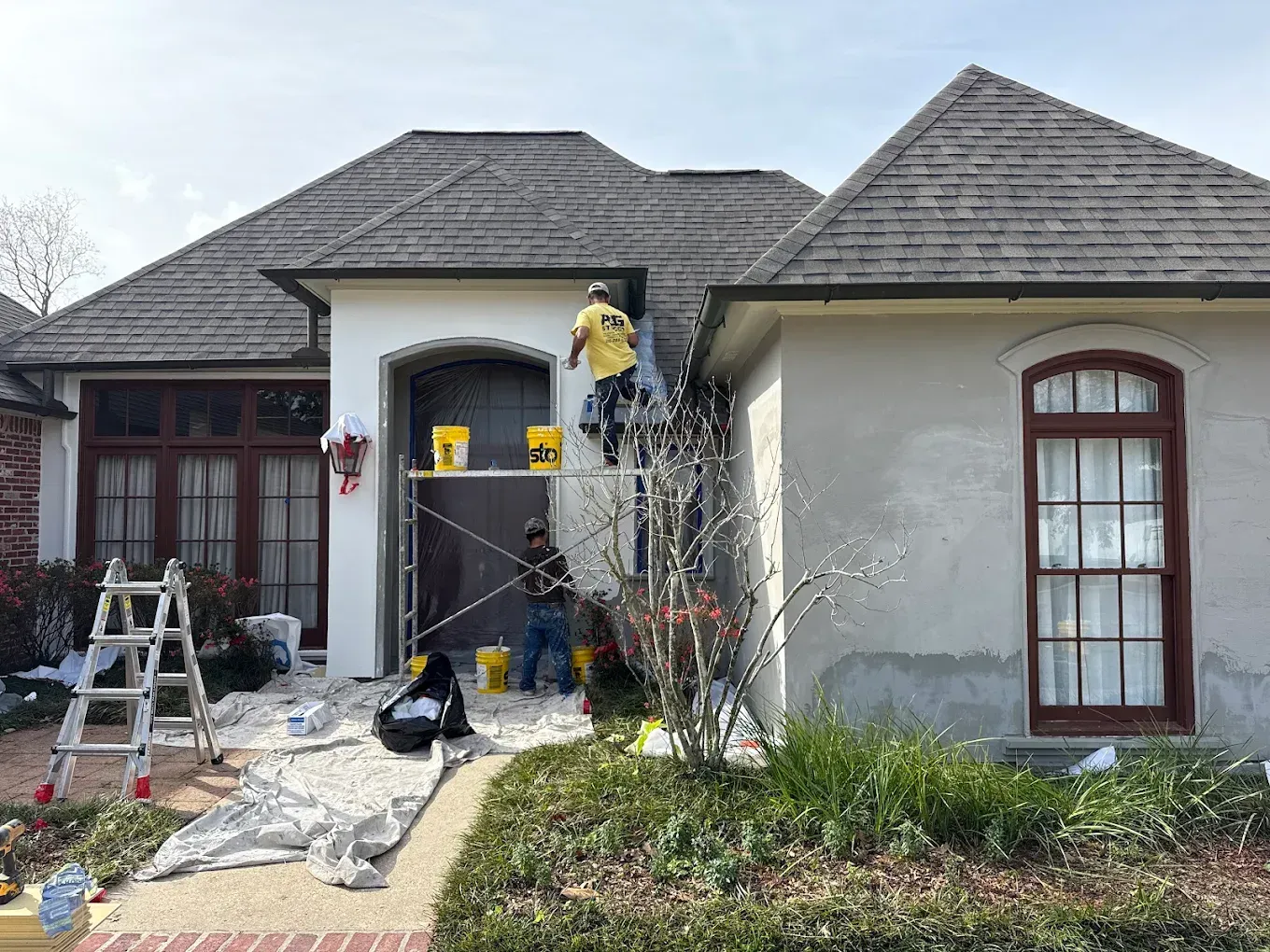 Workers painting a house. Two on ladders, applying stucco. Yellow buckets, brown trim, green grass.