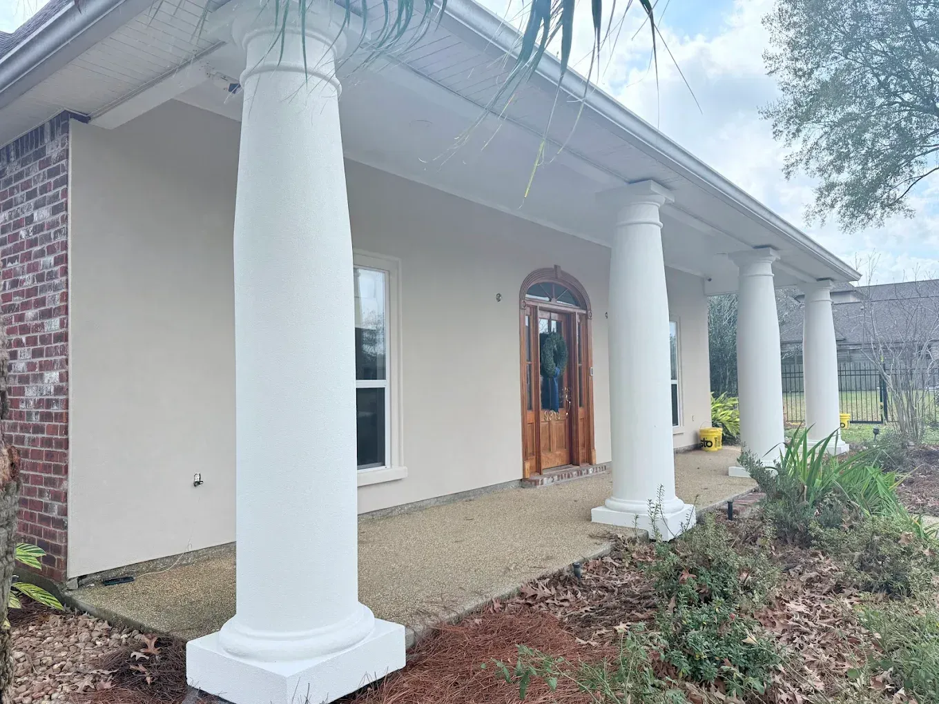 Exterior of a house with white columns, light stucco walls, and a wood door, under a cloudy sky.