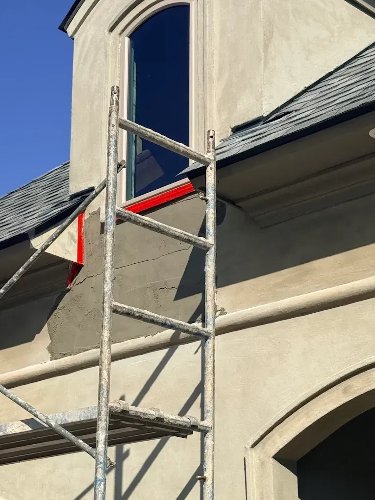 Scaffolding next to building with stucco walls, patching repairs, arched window and dark roof.