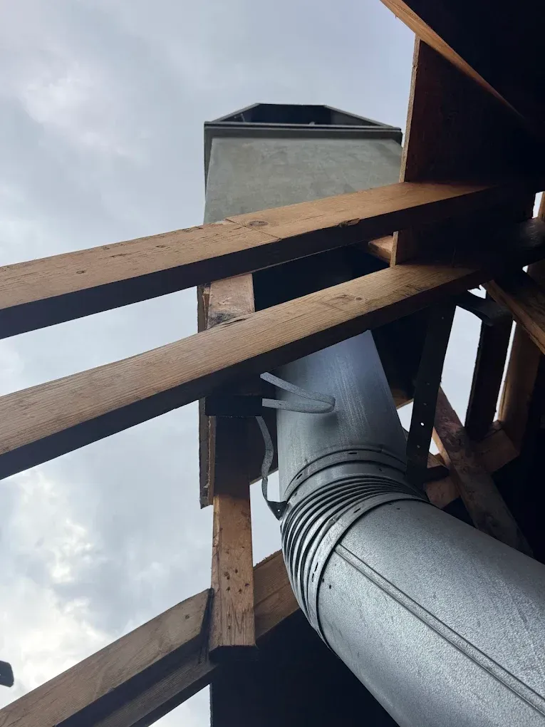 Chimney structure through a wooden attic frame with a metal pipe, against a cloudy sky.