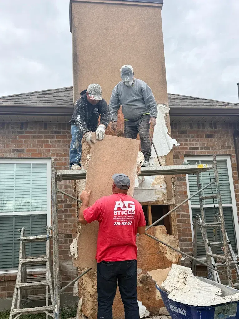 Three workers repair a chimney on a brick house; one holds a panel, two stand on scaffolding.