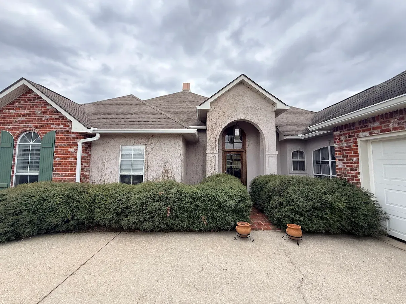 House exterior with brick and stucco, arched doorway, garage, and low green hedges. Overcast sky.