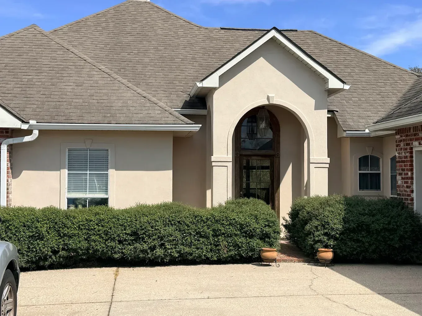 Beige stucco house with brown roof and arched entryway, green bushes in front.