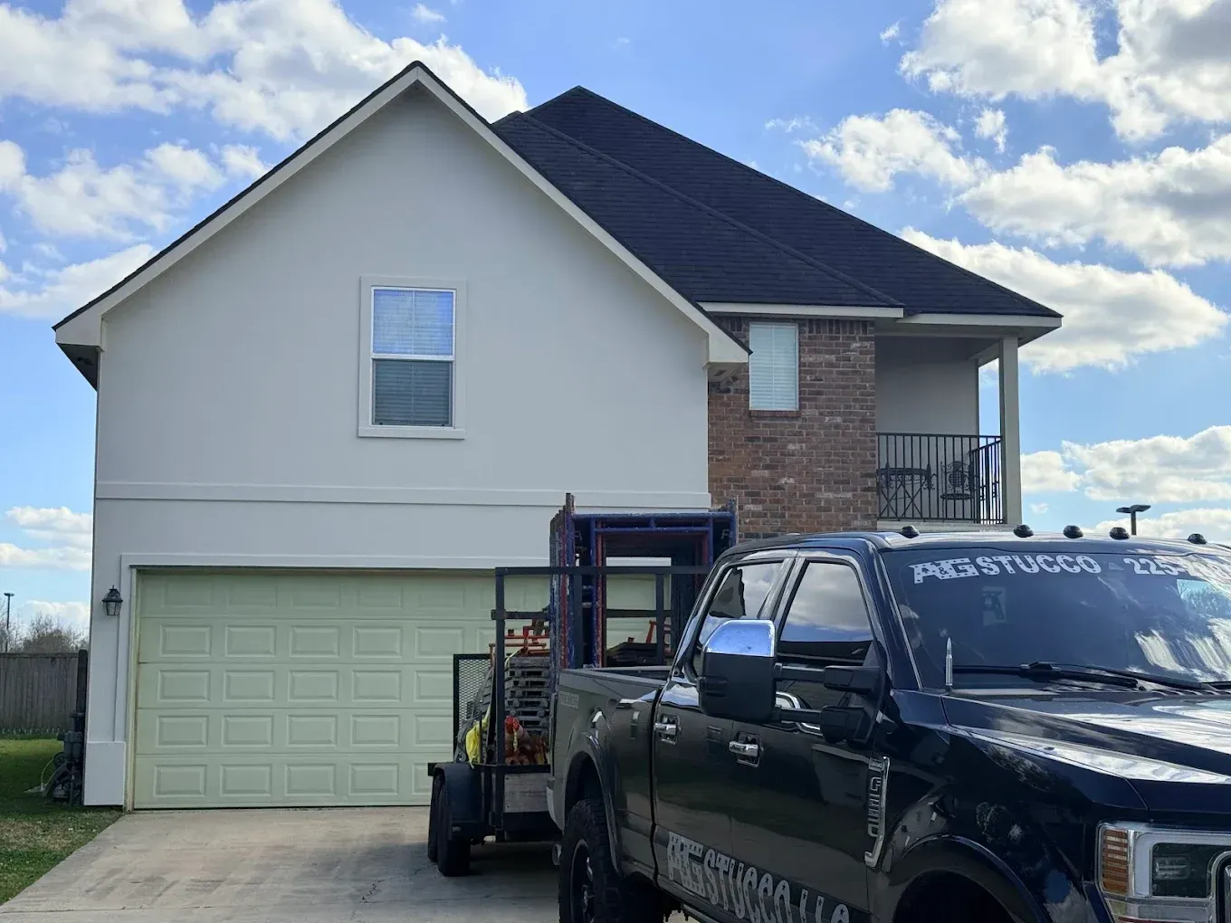 A two-story house with a black pickup truck and trailer in the driveway on a sunny day.