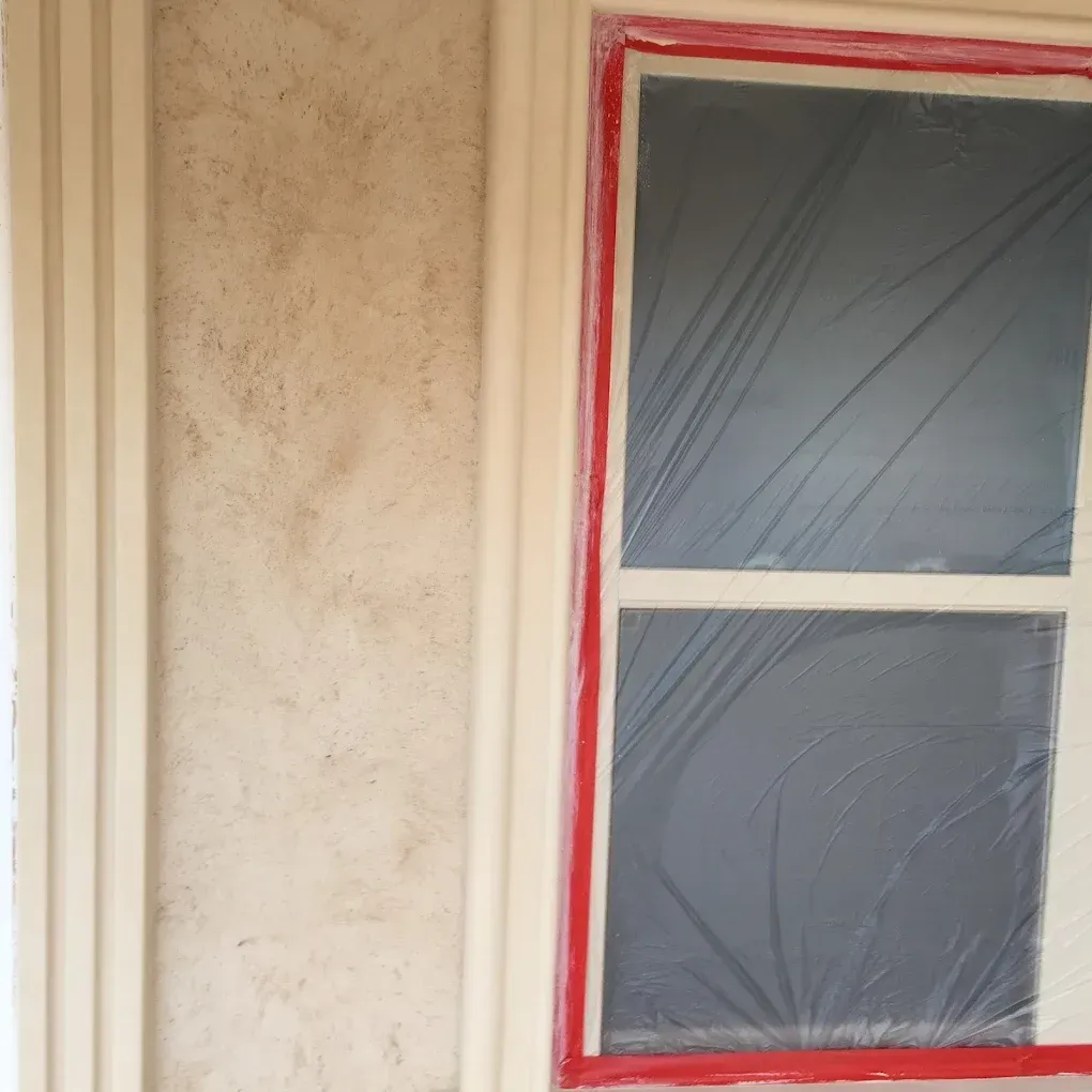 Exterior window with red tape, plastic covering, beige wall, and trim.