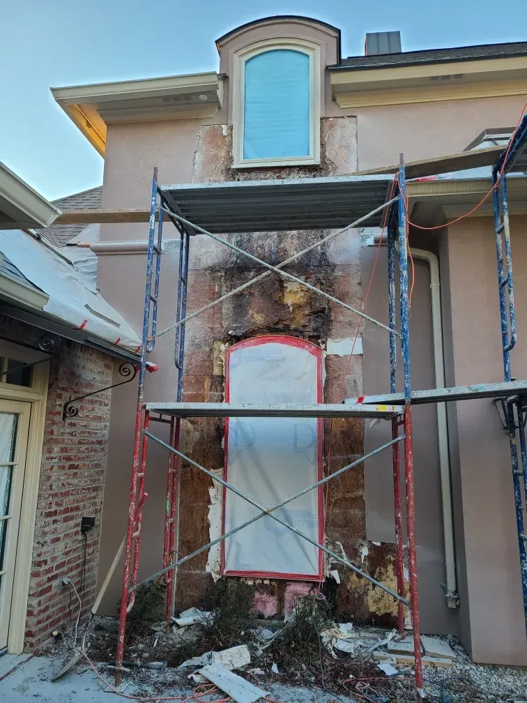 Scaffolding on a stucco wall undergoing repair, with a window above and exposed brick visible.