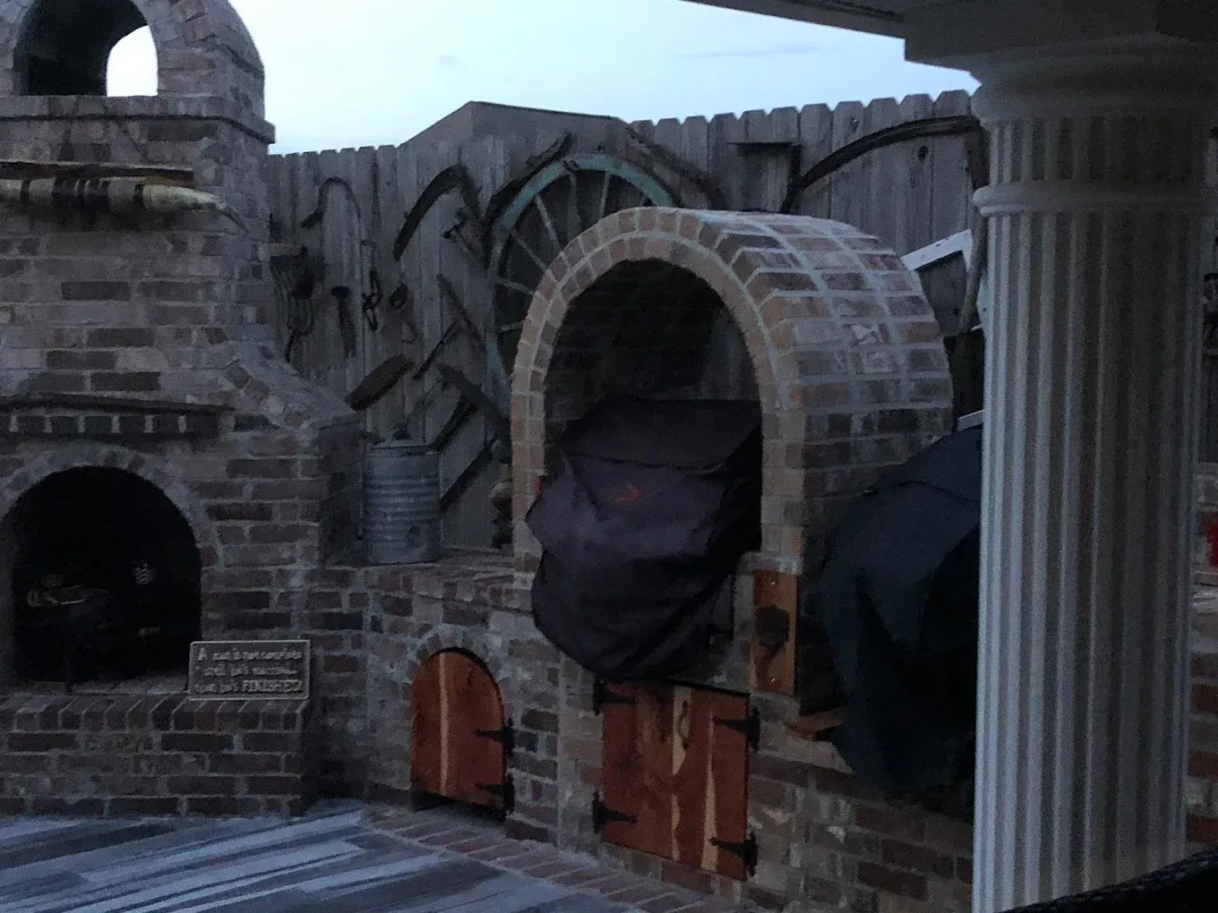 Brick structures with arched doorways and a water wheel against a wooden fence.