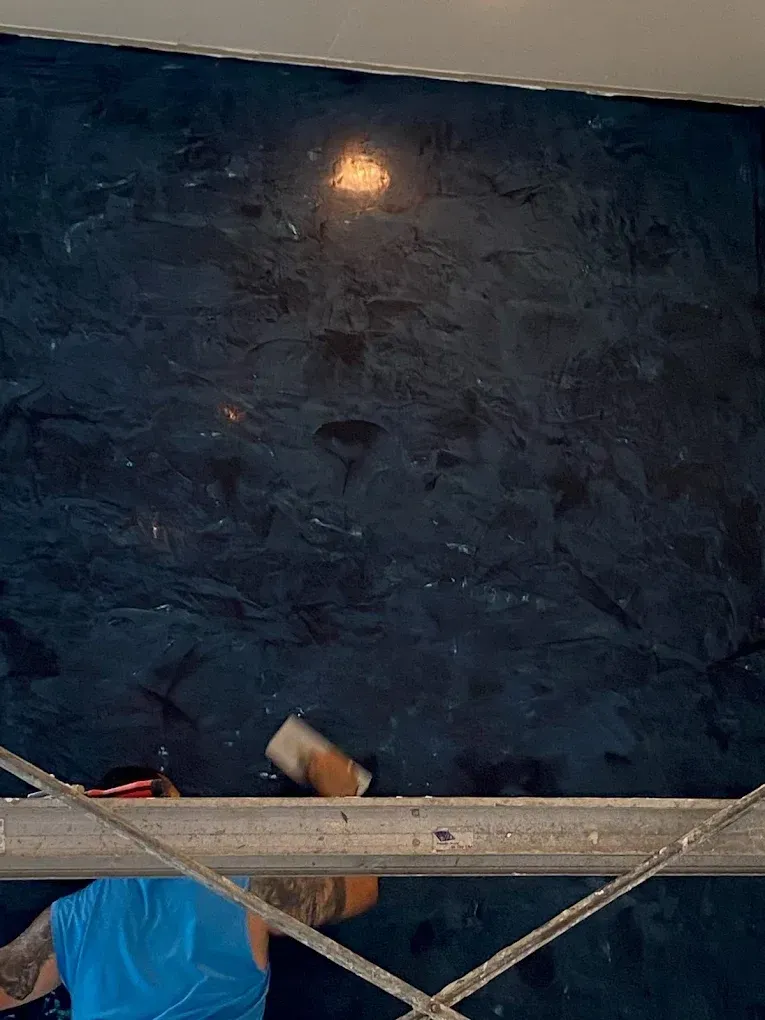 Person applying decorative finish to a dark blue wall from a ladder, with tools visible.
