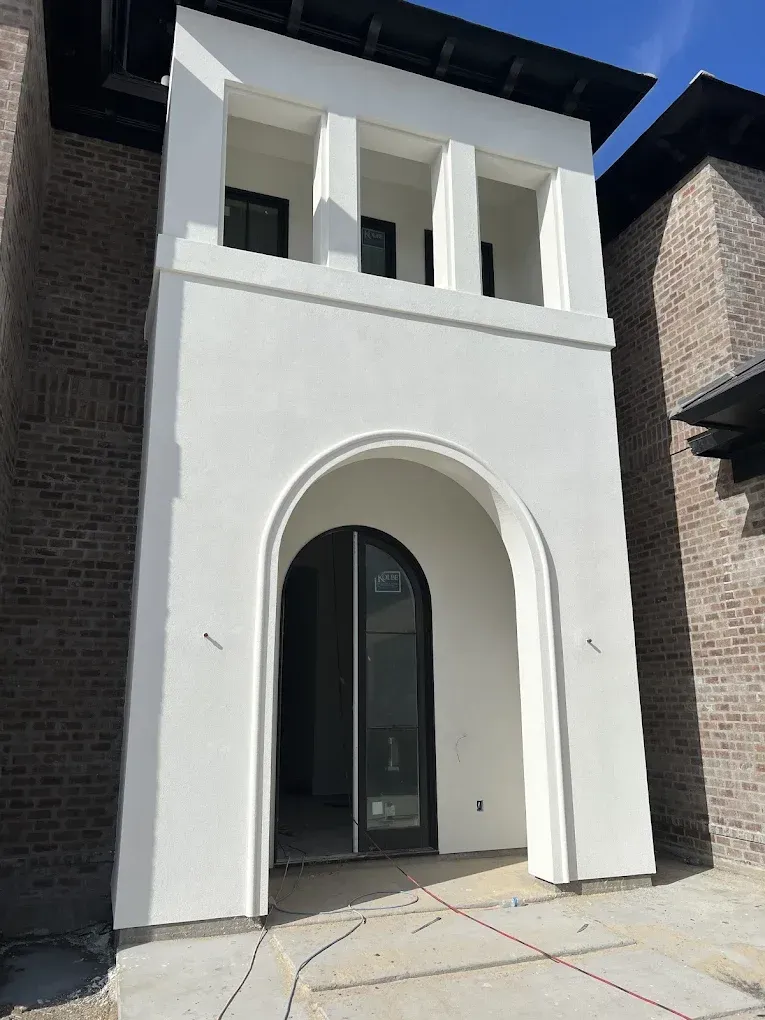 White stucco entrance with arched doorway and pillars, flanked by brick, under a dark roof.