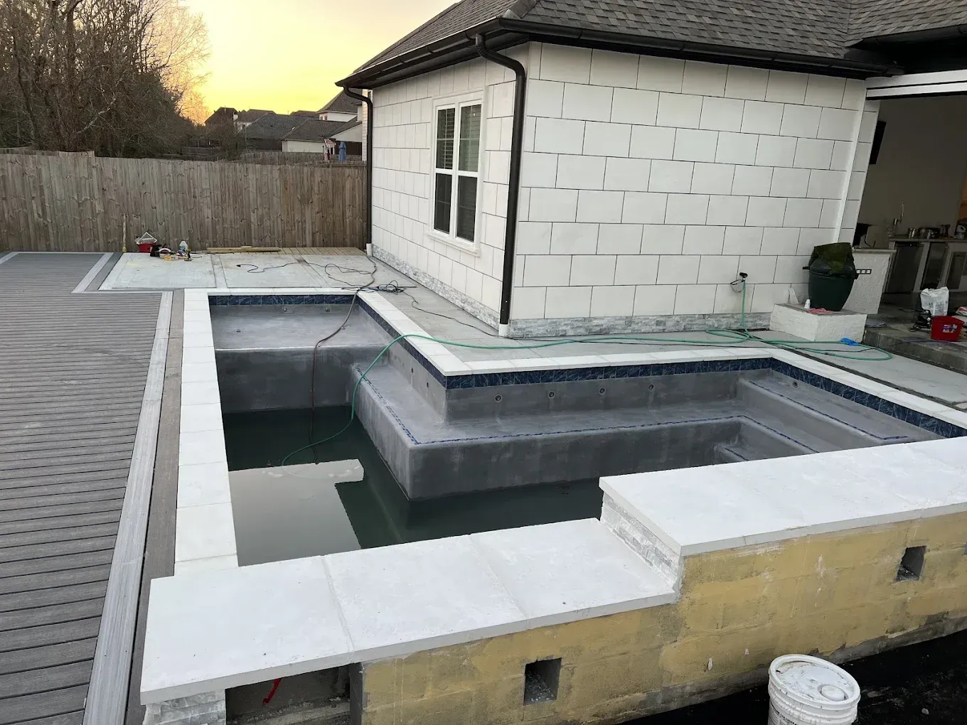 Pool with steps, light-colored tile surround, adjacent to a building with textured white siding and a wood fence.