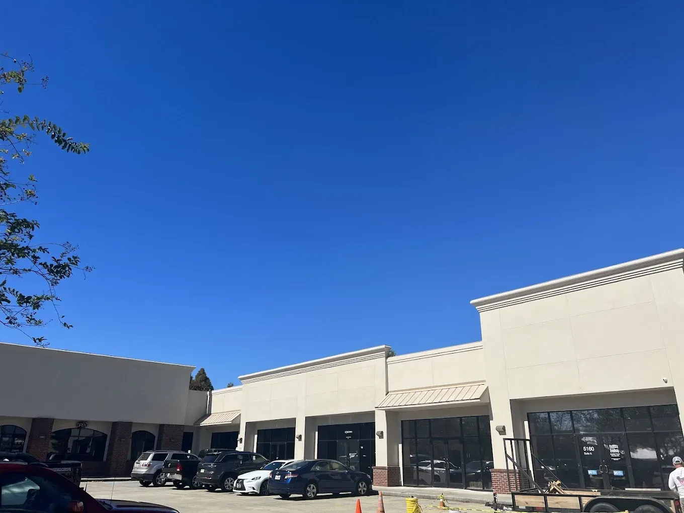 Shops under bright blue sky, with cars parked out front. Building is light-colored with dark windows.