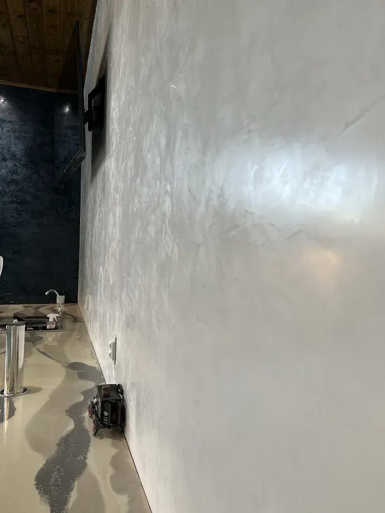 White Venetian plaster wall reflects light. Bathroom with sink, toilet, dark accent wall, and wood ceiling in background.