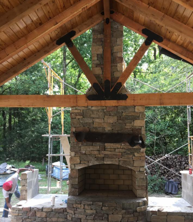 A stone fireplace under a timber-framed pavilion roof with decorative black metal bracing and a person working nearby.