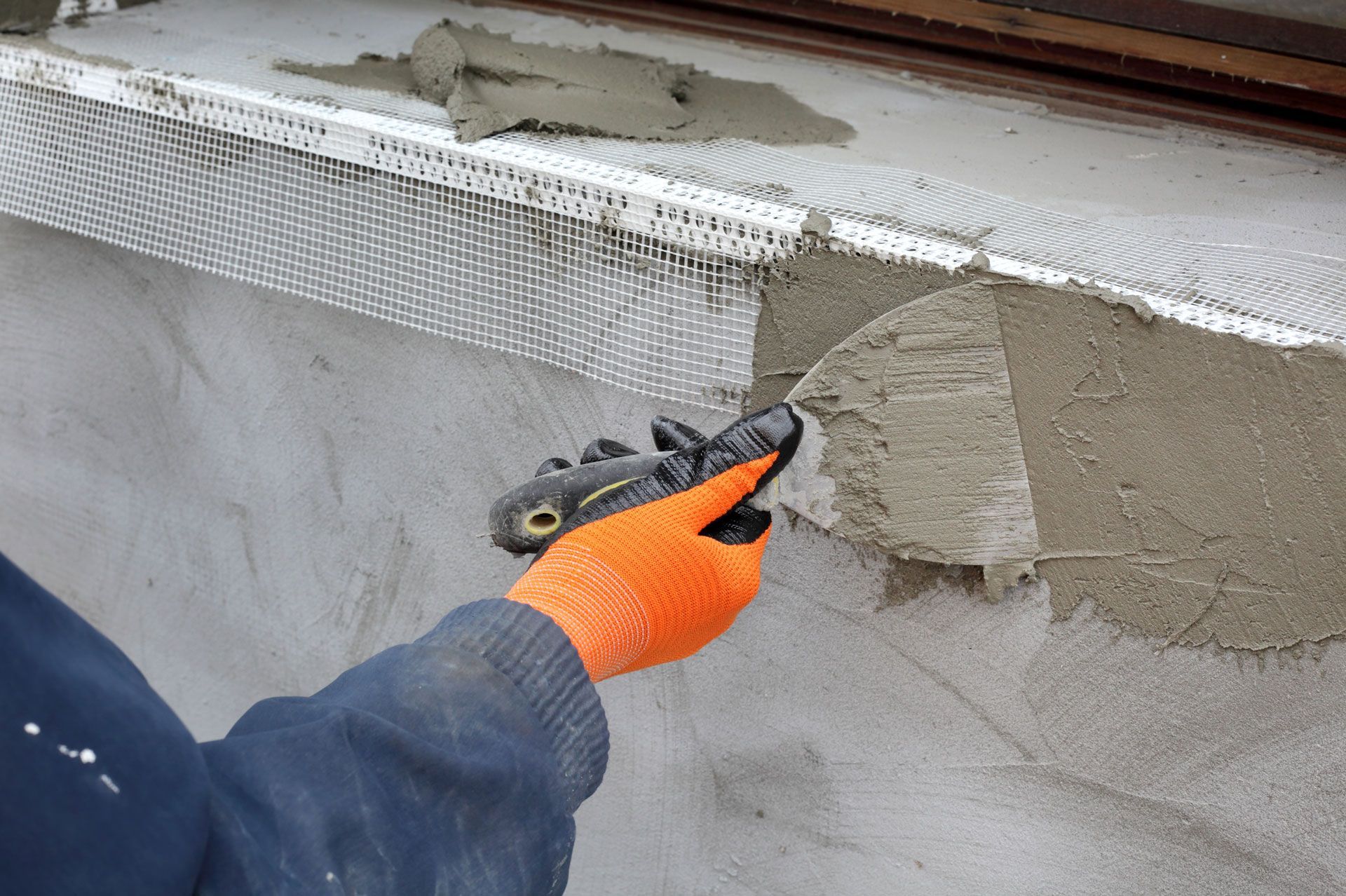 Person wearing orange gloves, applying cement to a wall with a trowel; metal mesh embedded.