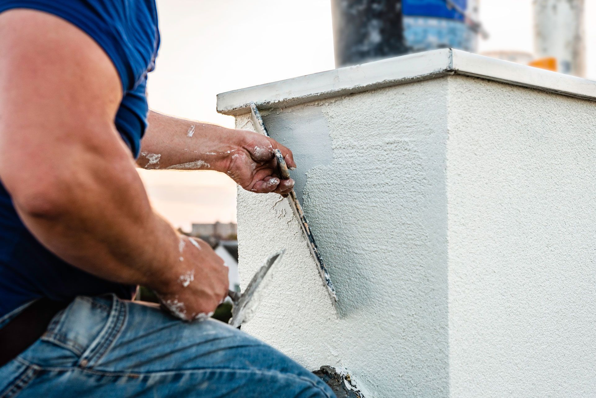 A person in jeans and a blue shirt applies stucco to a white wall using a trowel.