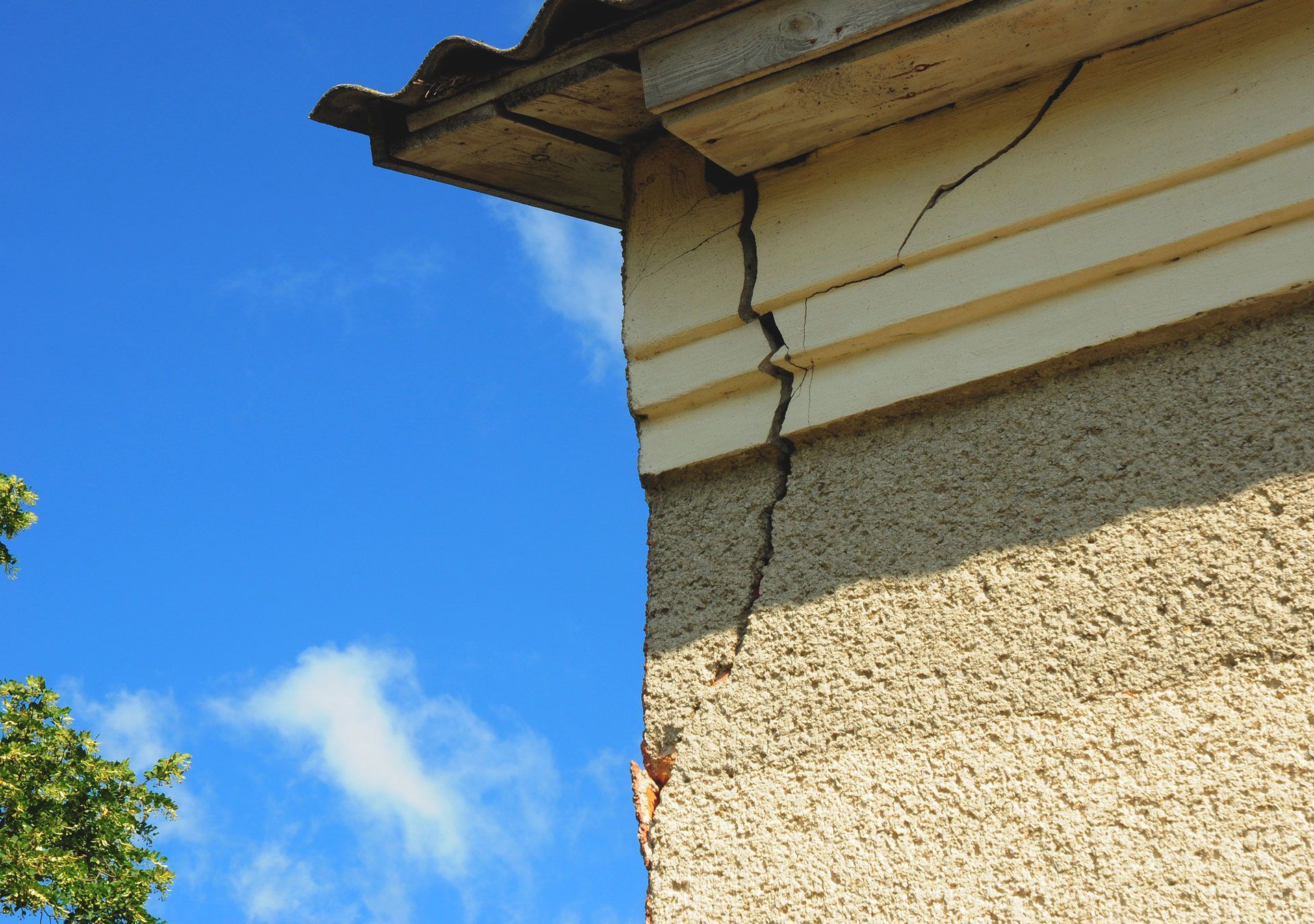 Exterior corner of a building with a prominent vertical crack in the stucco and siding. Blue sky in background.