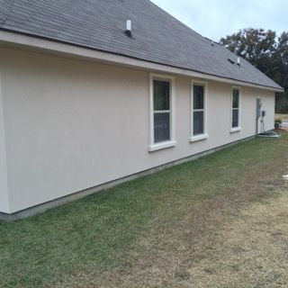 A white house with a gray roof and a lot of windows is sitting on top of a lush green field.