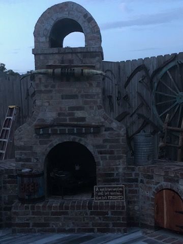 A rustic outdoor brick fireplace decorated with longhorns and antique farm tools against a wooden fence.