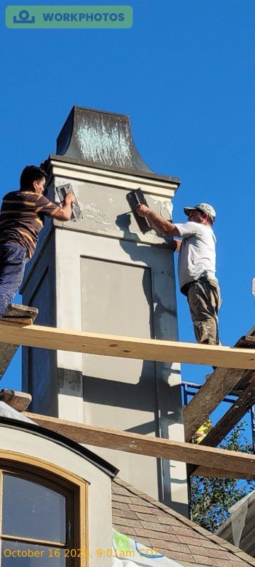Two workers on scaffolding apply plaster to the exterior of a chimney against a clear blue sky.