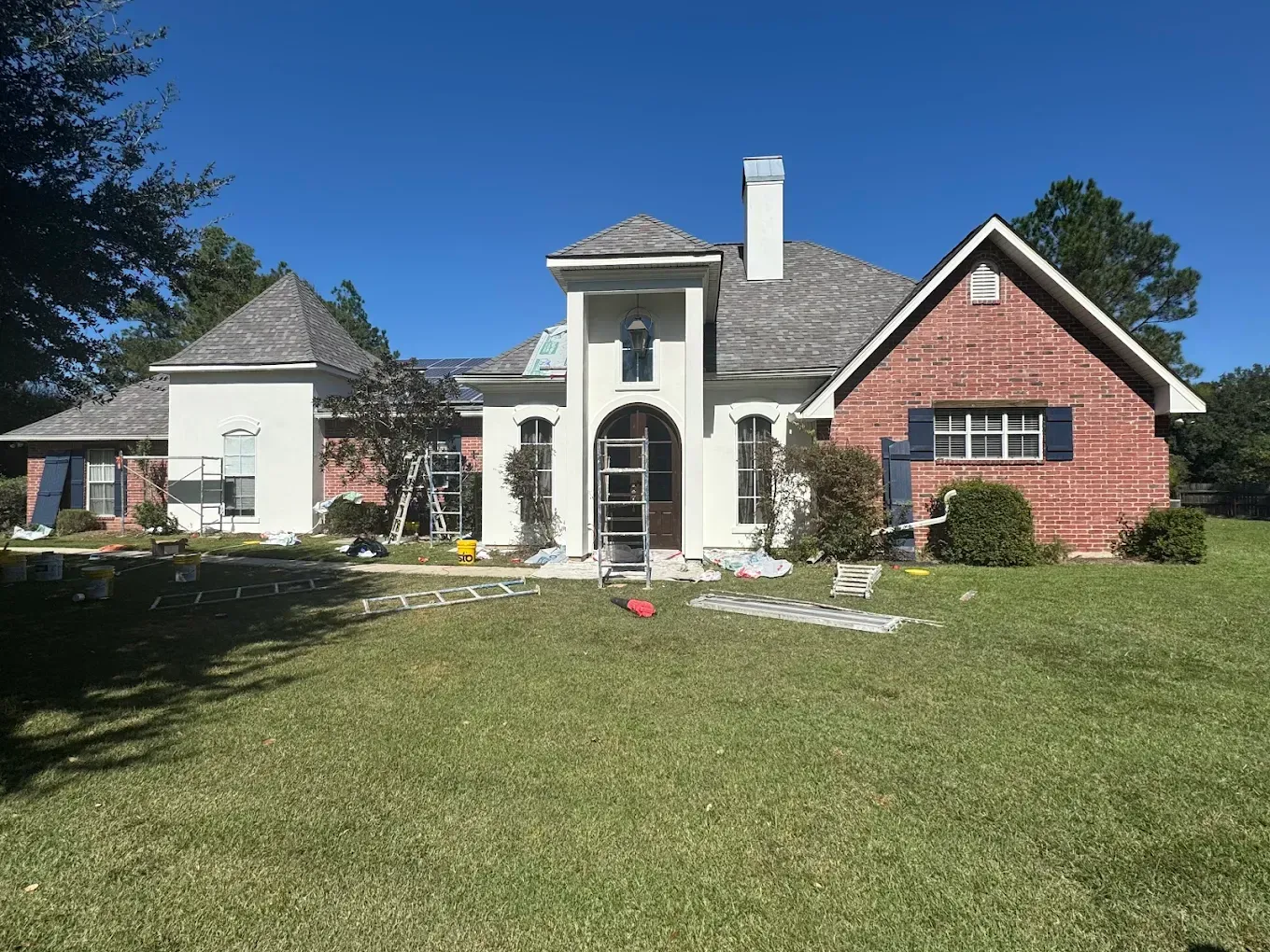 House exterior with brick, white stucco, and cedar shake. Ladders and painting supplies are visible.