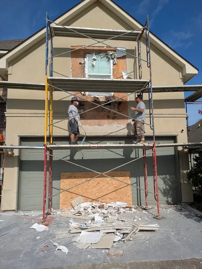 Men on scaffolding repairing stucco wall around a window above a garage. Debris on ground.