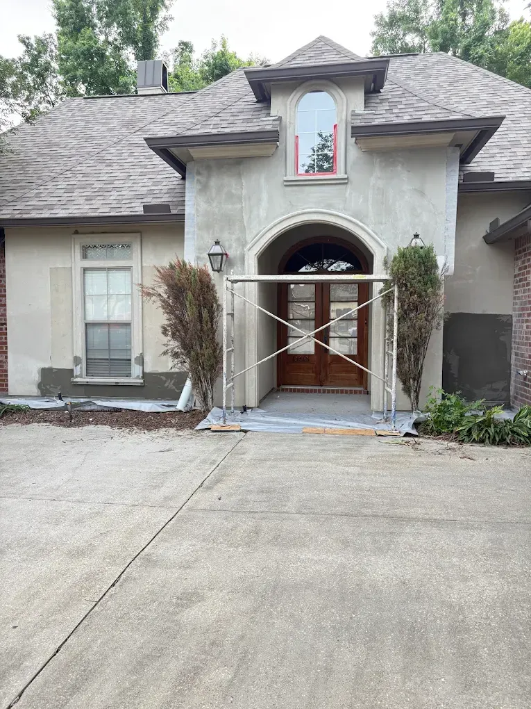 House exterior during renovation. Gray stucco and brick facade with scaffolding by the front door.