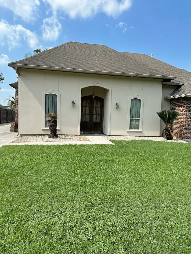 Beige house with arched doorway and brown roof, green lawn, blue sky.