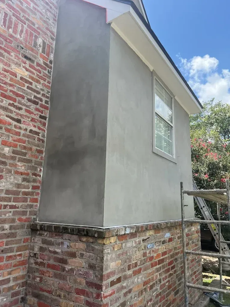 Brick chimney with gray stucco, beside a building with a window and white trim.