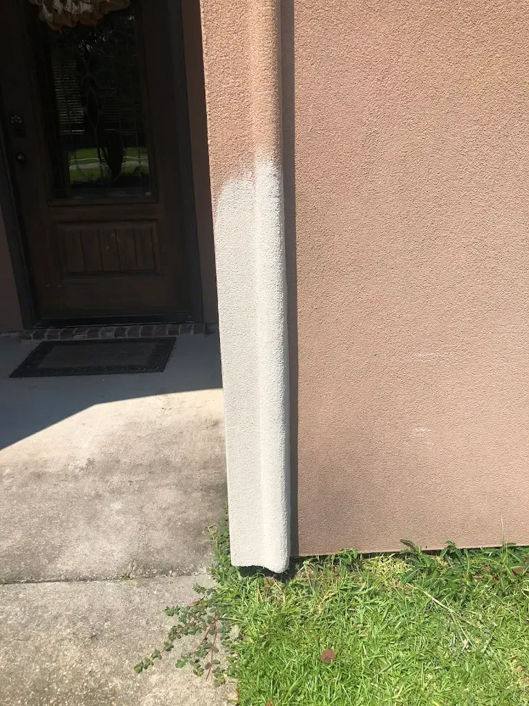 Exterior house corner with brown stucco wall and concrete pillar, near a grassy lawn and walkway.