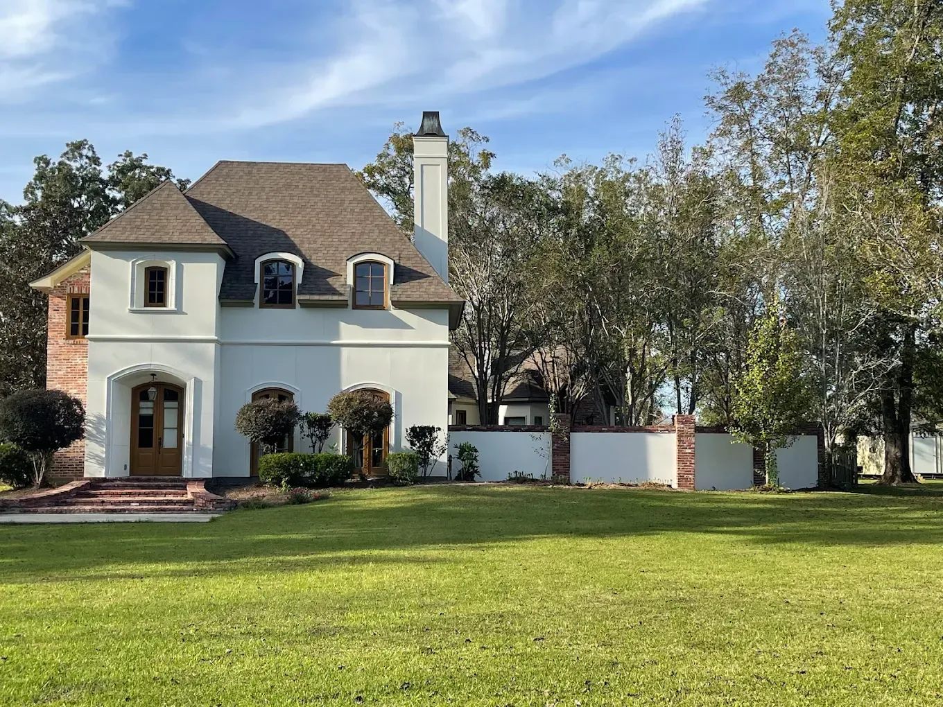 Two-story pale green house with a brown roof and a brick facade, set on a green lawn with a white fence.