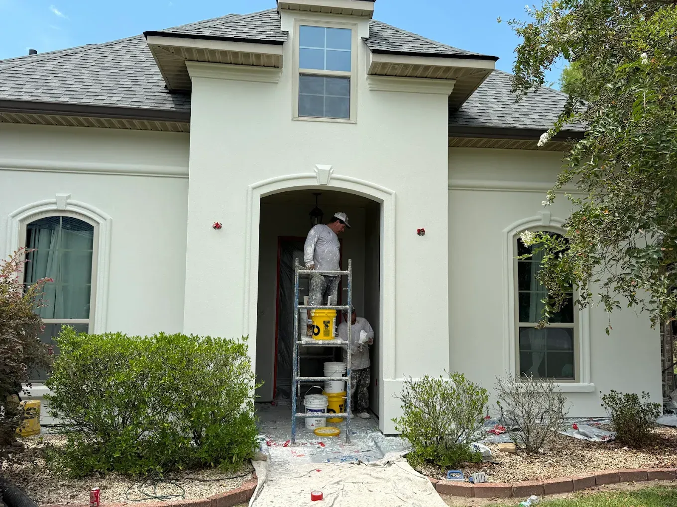 Workers painting a white house exterior with arched entryway. One stands on a ladder. Green bushes in front.