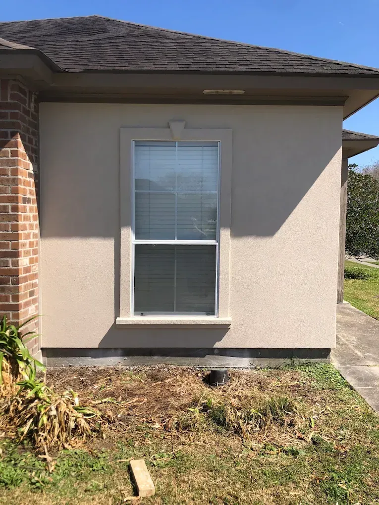 Tan stucco exterior wall with a window, brick corner, and brown roof under a blue sky.
