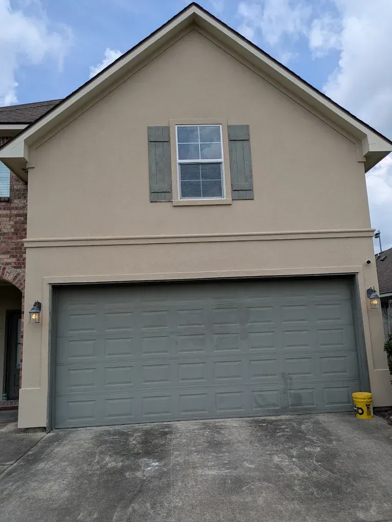Tan two-story house with gray garage door, window with shutters, and cloudy sky.